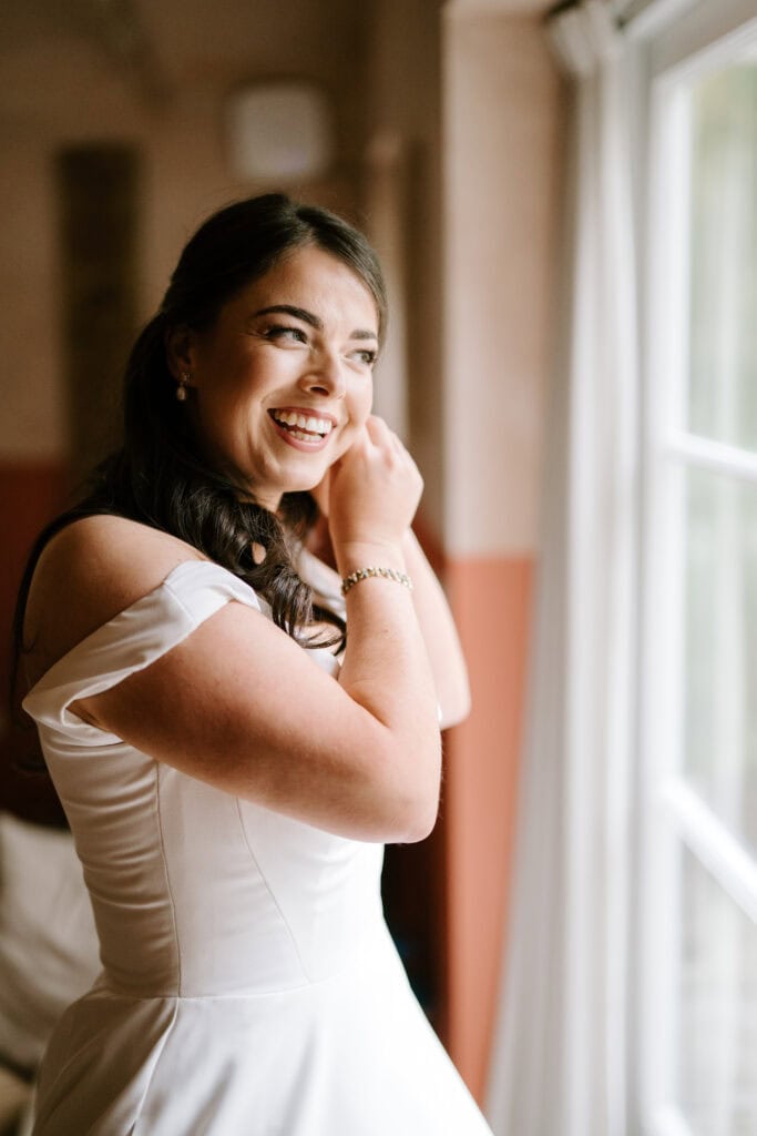 Smiling bride adjusts earring by window.