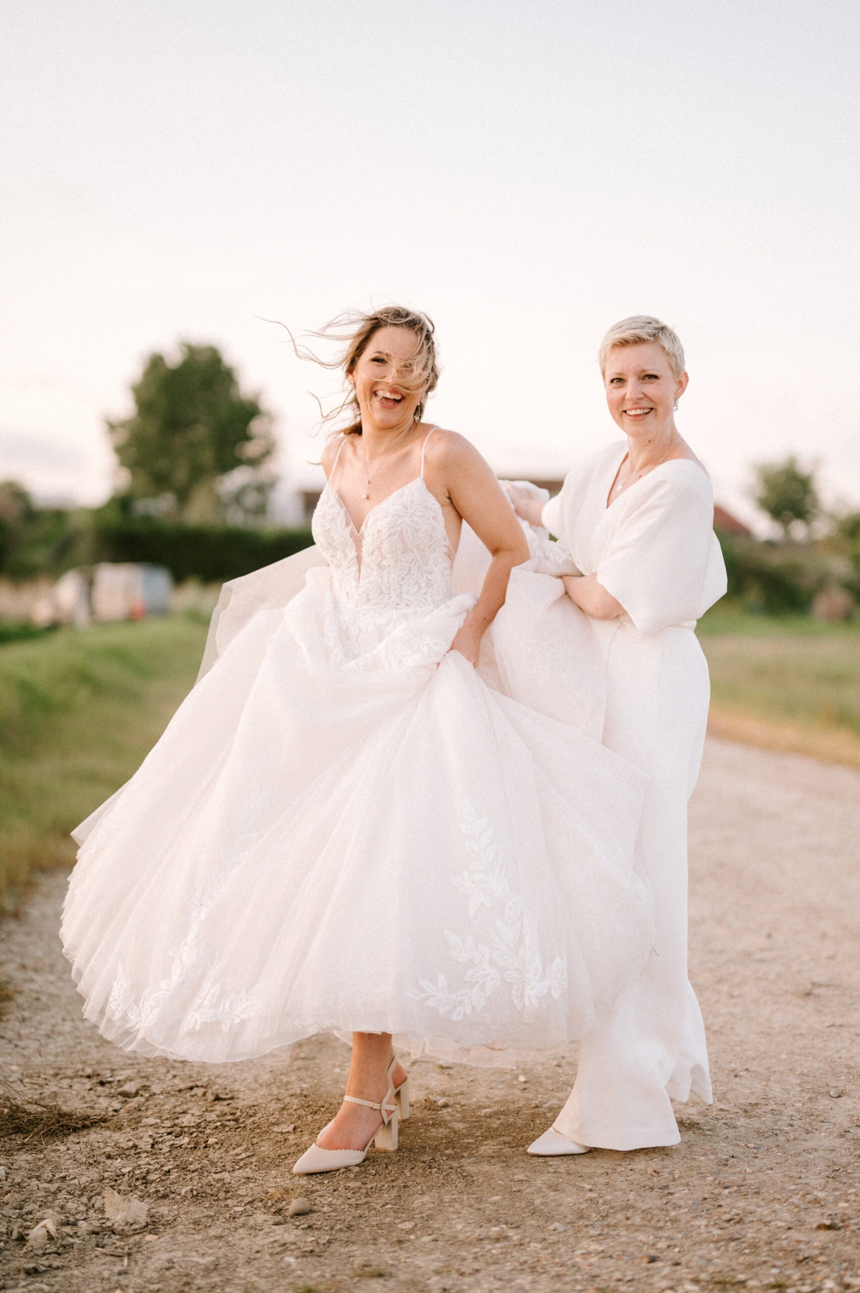 Two smiling brides in elegant dresses outdoors.
